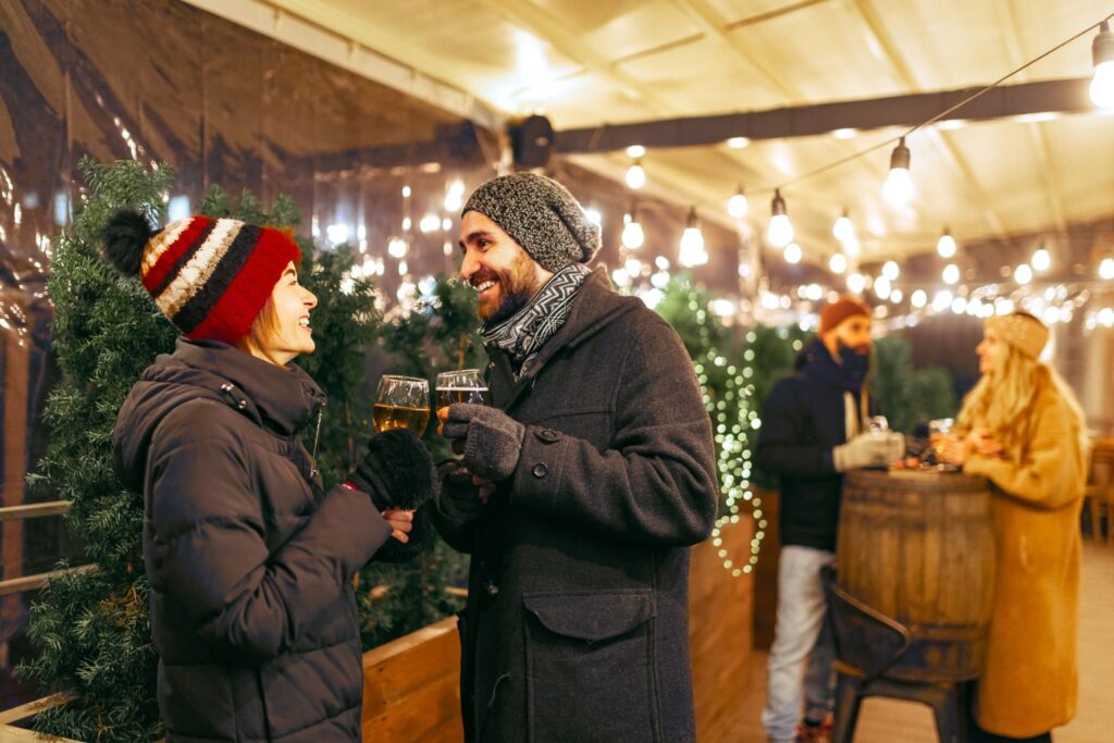 A couple smiling at each other at a German christmas market, they are wearing a warm jacket, scarf and hats as well as gloves, those items should be on any packing list for Germany in winter