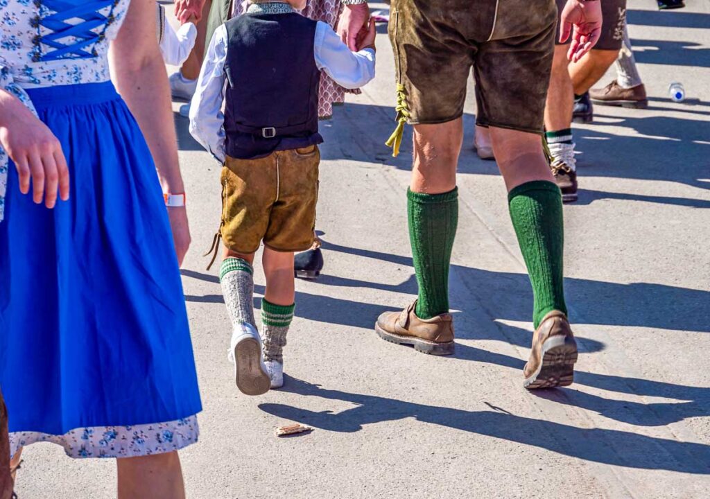 A young boy dressed in traditional Oktoberfest clothing, wearing mini lederhosen, knee-high socks, and a vest, walking hand-in-hand with adults in Bavarian attire at the Oktoberfest festival in Munich.