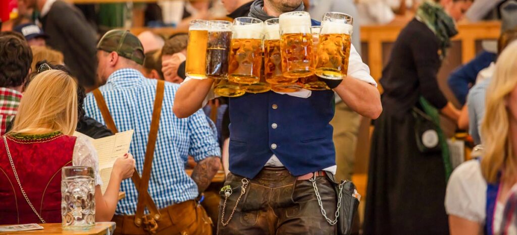 A waiter in a blue vest and leather pants skillfully carrying multiple large beer mugs filled with golden beer and topped with frothy foam at a crowded Oktoberfest beer hall in Munich.