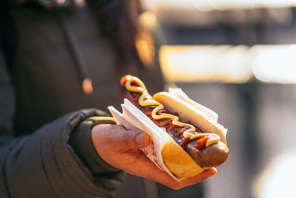 Close-up of a hand holding a Bratwurst in a bun, topped with ketchup and mustard—popular Oktoberfest food enjoyed at outdoor stands.