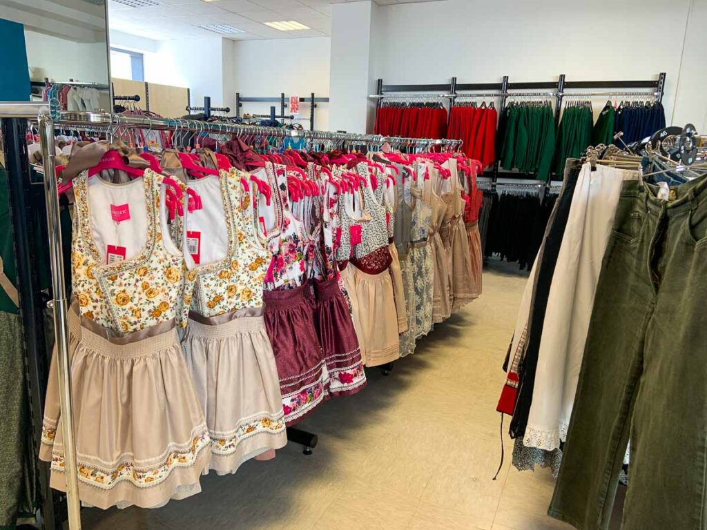 Dirndl dresses in a store – A variety of colorful dirndl dresses with floral patterns and lace trims hanging neatly on racks in a store. The dresses have bright pink hangers, and traditional clothing can be seen in the background. This is a great option for visitors searching where to buy a dirndl in Munich before Oktoberfest.