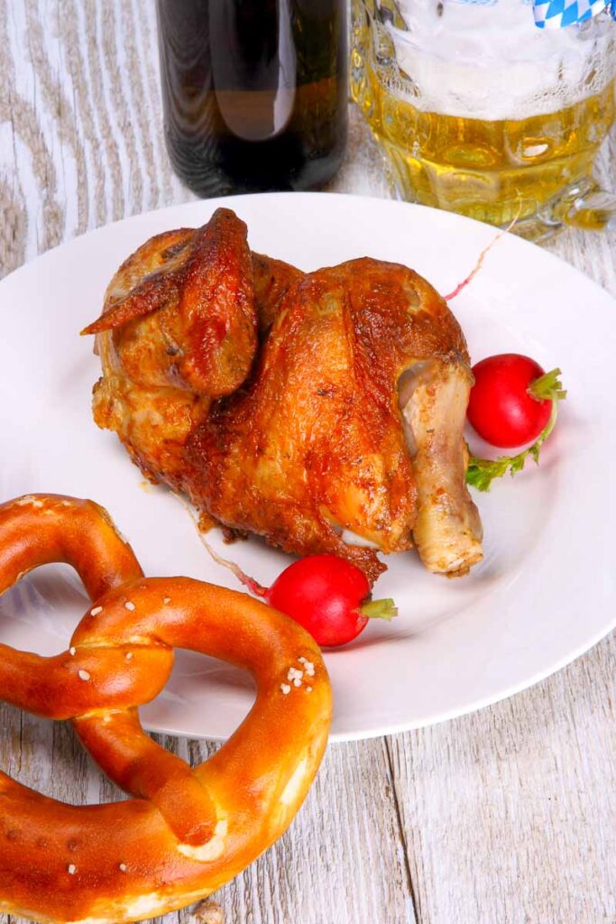 Plate of crispy roasted half chicken (Hendl) served with red radishes and a fresh pretzel, accompanied by a glass of beer—traditional Oktoberfest food.