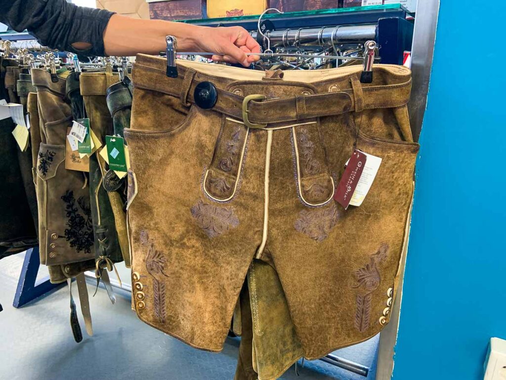 Traditional lederhosen on display – A person holds up a pair of brown suede lederhosen with decorative embroidery and metal buttons at the hem. More lederhosen hang in the background on a clothing rack. If you're wondering where to buy a dirndl in Munich, many second-hand and specialty stores also carry lederhosen.