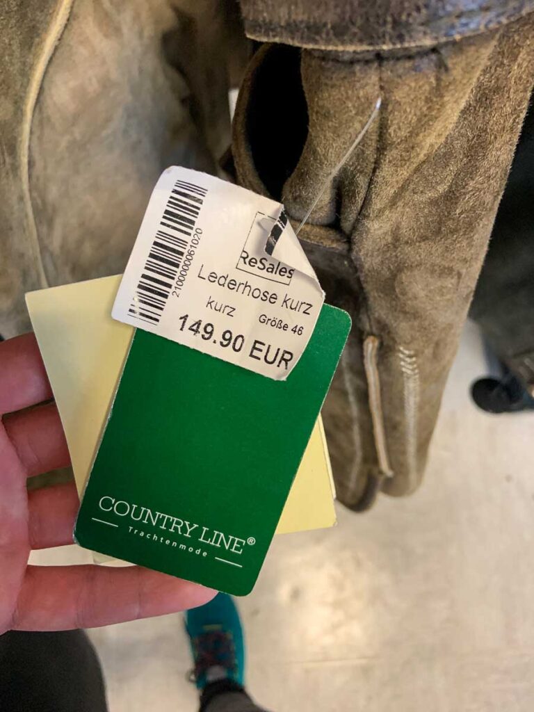 Close-up of a price tag on lederhosen – A person holding a green price tag for short lederhosen from the brand "Country Line," priced at €149.90. The lederhosen are visible in the background, hanging on a rack. A great find for those searching for where to buy a dirndl in Munich or traditional Bavarian clothing.