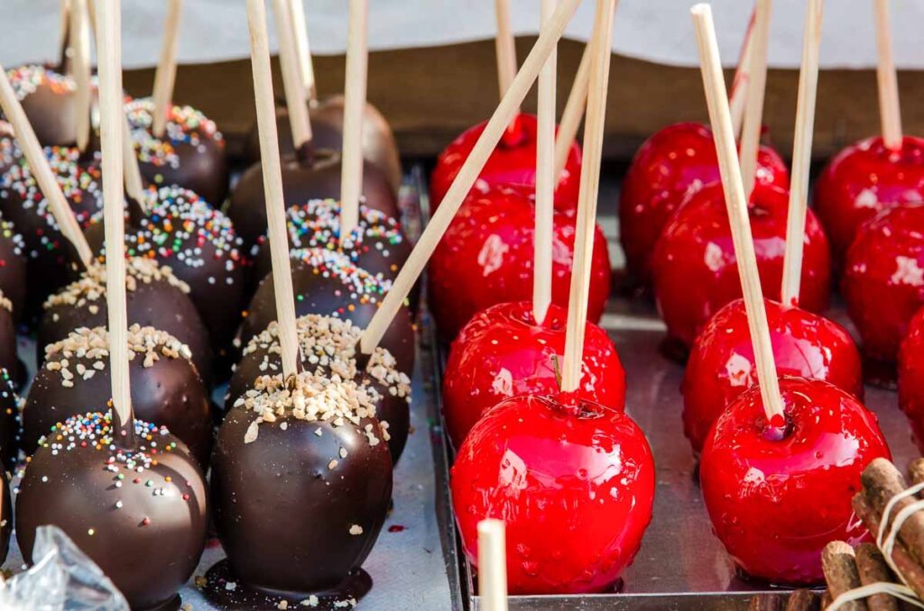Close-up of chocolate-dipped and glossy red candy apples with wooden sticks, topped with sprinkles and nuts—popular sweet treats at Oktoberfest desserts.