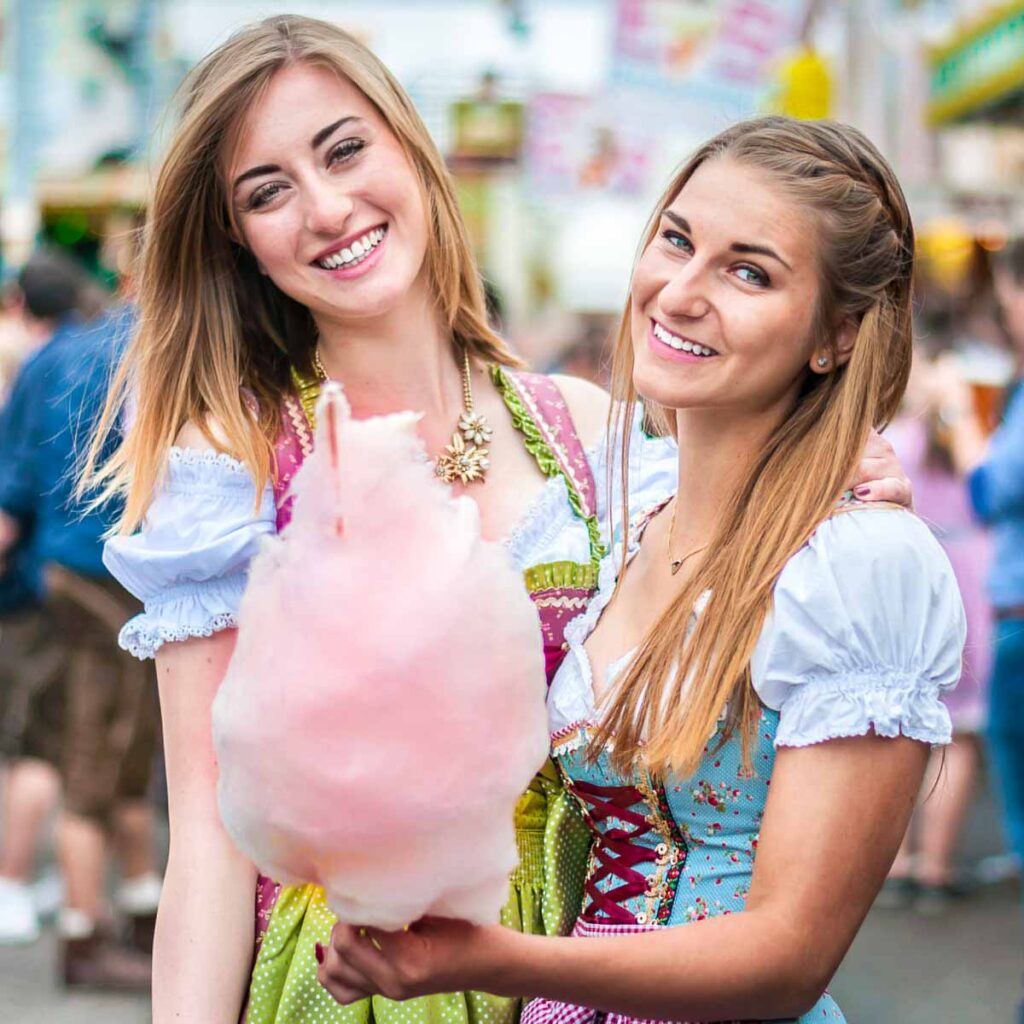 Two smiling women dressed in traditional Dirndl dresses holding pink cotton candy at Oktoberfest in Munich —combining festive attire with fun fair-style Oktoberfest food.