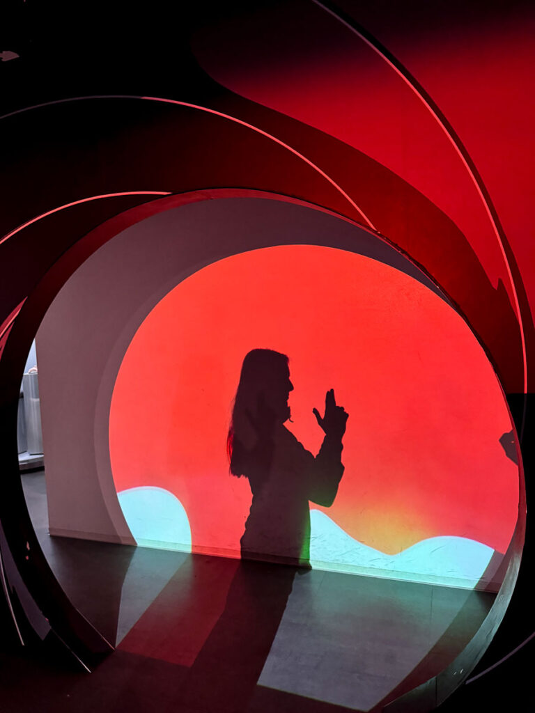 A person strikes a classic finger-gun spy pose in front of a red and black backdrop resembling the iconic James Bond gun barrel design. A fun photo op featured in many spy museum Berlin reviews.