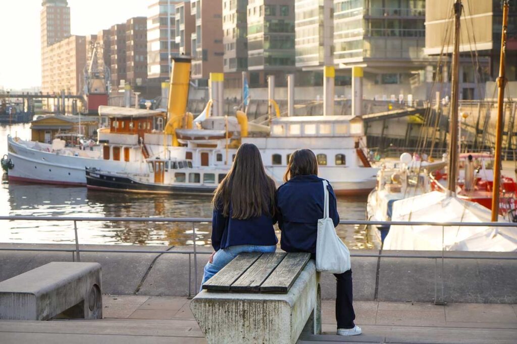 Two women sit on a bench by the canal in Hamburg's Speicherstadt district, with historic ships and modern buildings reflecting the city's maritime charm.