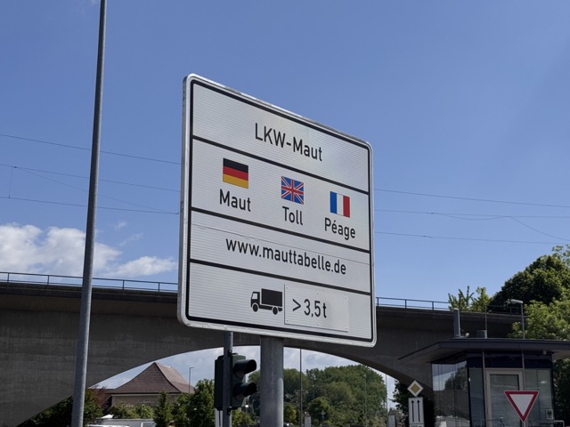 A road sign in Germany displaying “LKW-Maut” with German, British, and French flags next to the words “Maut,” “Toll,” and “Péage.” Below is a URL “www.mauttabelle.de” and an icon of a truck over 3.5 tons, indicating a toll for heavy vehicles.
